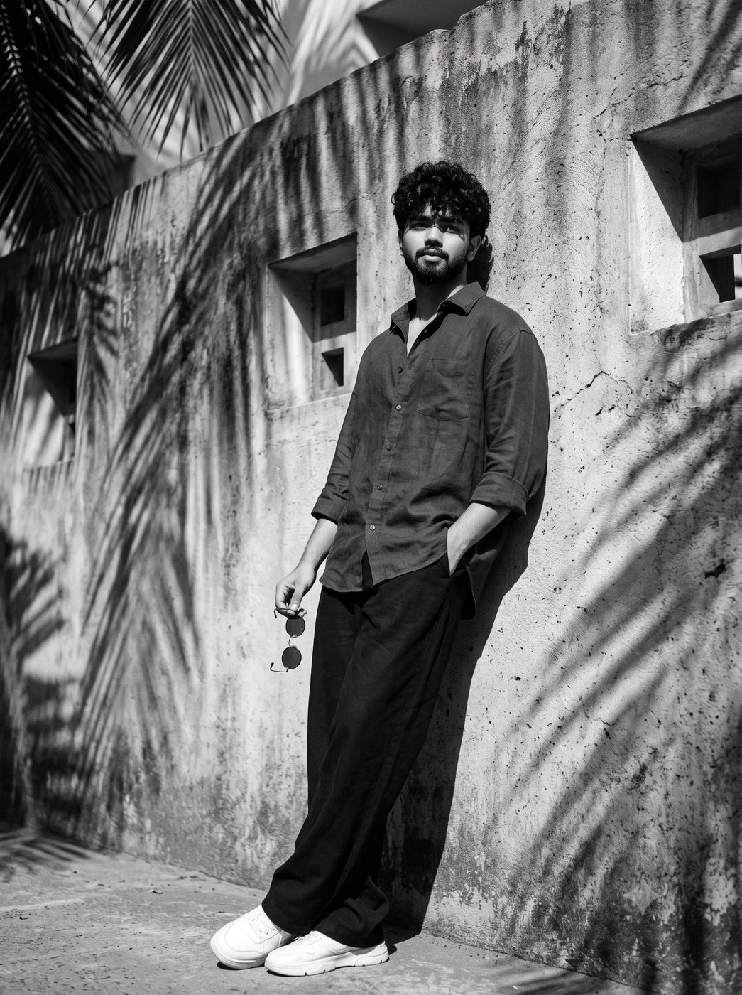 AI Photo Editing Prompt: Artistic B&W photography of a curly-haired man leaning elegantly on a rough wall with diagonal shadows and holding classic round sunglasses.