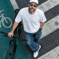 ChatGPT Prompt: ChatGPT generated Top-down aerial street photography of a bearded man in an oversized white t-shirt, baggy blue jeans, white cap, and reflective sunglasses holding a black bicycle on a zebra crossing next to a teal bike lane.