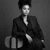 Professional Woman Studio Photography AI Prompt. AI-generated portrait of a woman in a pinstripe suit, with her curly hair up, leaning against a chair in black and white.