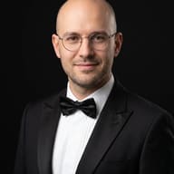 Man in Tuxedo Portrait AI Photo Editing Prompt. AI generated portrait of a bald man with round glasses wearing a black tuxedo, white pleated shirt, and black bow tie, against a black background.