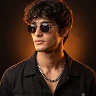 AI generated studio portrait of a young man with dark curly hair, wearing round sunglasses, a silver chain, and a black jacket, with warm orange rim light on a dark background.