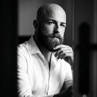 Gemini AI Prompt: Editorial B&W studio shot of a thoughtful bald man with a thick beard posing elegantly behind a softly blurred foreground silhouette.