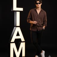 AI generated studio portrait of a dark-haired man in a brown shirt and black pants wearing sunglasses and holding a second pair, standing next to a large illuminated sign spelling out LIAM.