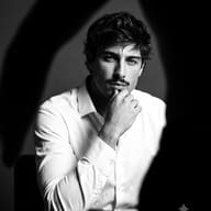 AI Photo Prompt: Dramatic monochrome fashion image of a striking man with a mustache in a white shirt framed by deep shadows and intense highlights.