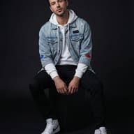 AI generated studio portrait of a man sitting on a black block against a black background, wearing a white hoodie, distressed denim jacket with patches, black jeans, and white Nike sneakers.