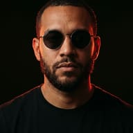 Man Red Lighting Portrait AI Photo Editing Prompt. AI generated studio portrait of a bearded man wearing round sunglasses and a black t-shirt illuminated by dramatic red rim lighting against a dark background.