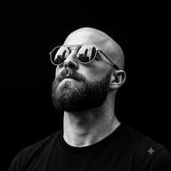 AI Prompt: High-contrast black and white portrait of a bald man with a beard gazing upward, wearing round sunglasses reflecting a city skyline.