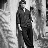 AI Image Prompt: Premium monochrome lifestyle image of a stylish guy in a linen shirt and chunky sneakers posing near a wall with small square windows.