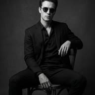 Man in Dark Suit B&W Portrait AI Photo Editing Prompt. AI generated black and white studio portrait of a young man wearing a suit and round sunglasses sitting on a wooden chair.