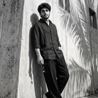 AI Prompt Prompt: Striking minimal portrait of a confident bearded man in dark clothing standing casually against a sunlit wall with palm frond shadows.