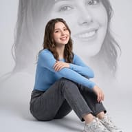 Double Exposure Portrait AI Photo Editing Prompt. AI generated studio portrait of a smiling young woman in a blue shirt and grey jeans sitting on the floor with a large greyscale background face overlay.