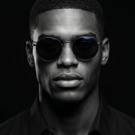 AI generated studio portrait of a sharp-featured man with a buzz cut and round sunglasses in dramatic dark lighting.