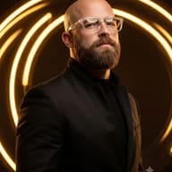 AI Images Generation Prompt: Dramatic low-key portrait of a bald man with a full beard wearing clear eyeglasses and a black suit jacket, standing in front of a warm golden neon ring light.