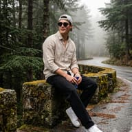 Travel Portrait of Guy on Misty Forest Road AI Photo Editing Prompt. AI generated outdoor portrait of a young man in a cap and sunglasses sitting on a mossy stone wall beside a wet mountain road in a foggy pine forest.