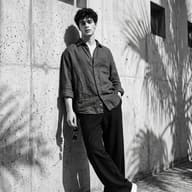AI Photo Editing Prompt: Classic fashion editorial portrait showing a handsome young man standing casually with one hand in his pocket against a shadow-patterned wall.