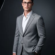 AI Image Prompt: Cinematic corporate photo of a young man with curly hair wearing dark glasses and a light grey suit posing confidently against a dark background.