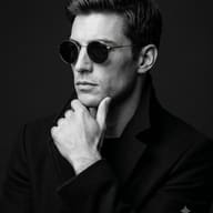 Man in Sunglasses B&W Portrait AI Photo Editing Prompt. AI generated studio portrait of a stylish man wearing a high-collar coat and round sunglasses with his hand on his chin.