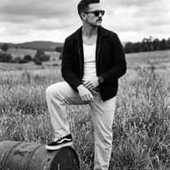 Stylish Man on Rusty Barrel in Black and White AI Photo Editing Prompt. AI generated black and white outdoor portrait of a stylish man in sunglasses and a casual jacket resting his foot on a rusty barrel in a grassy field.