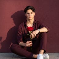 Gemini AI generated photo of a Young man with dark wavy hair in a burgundy shirt and trousers sitting on gray stone pavement holding a single red rose against a burgundy textured wall