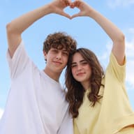 AI generated outdoor portrait photograph of a young smiling couple making a heart shape with their hands against a clear blue sky.