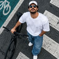 AI Photo Prompt: High-angle urban fashion portrait of a confident young man looking up at the camera while standing with a black bicycle on a textured asphalt road crossing with white stripes and a green cycle track.