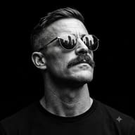 AI Photo Prompt: Chiaroscuro style B&W portrait of a man with a mustache in a black t-shirt, looking up with skyline reflections in his round sunglasses.