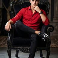 Gemini Ai Generated Image: A young man with wavy brown hair sits confidently in a luxurious, ornate black chair. He is dressed in a bright red button-down shirt, black skinny jeans, and black-and-white sneakers. With one leg crossed over the other and his hand resting thoughtfully on his chin, he gazes directly at the camera against a dark, elegant background.