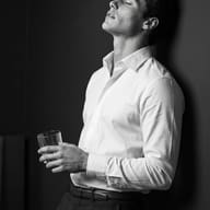 AI Generated Black and white hyper-realistic portrait of a young man in a crisp white button-down shirt leaning against a dark wall with his eyes closed, holding a rocks glass in cinematic chiaroscuro lighting.