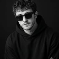 Monochrome Man Portrait AI Photo Editing Prompt. AI-generated high-contrast studio portrait of a young man with curly hair wearing dark rectangular sunglasses and a black hoodie, set against a dark background.