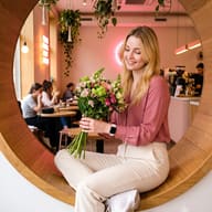 AI generated portrait of a blonde woman in a pink blouse holding flowers while sitting in a round wooden window seat in a cafe.