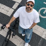 ChatGPT-generated realistic overhead street-style shot of a bearded man wearing a white cap, aviator sunglasses, a white T-shirt, and loose denim jeans, holding a road bike handle on a city crosswalk.