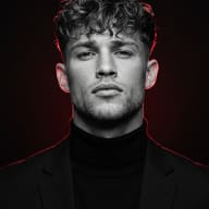 AI Generated dramatic, symmetrical studio portrait of a young man with wavy, curly hair and a serious, direct gaze, wearing a black blazer and turtleneck, with an intense, vibrant red rim light tracing his entire outline against a deep black background.