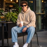 Cute Man with Coffee and Wide-Leg Jeans Seated Lifestyle AI Photo Editing Prompt. AI generated photorealistic portrait of a smiling young man wearing sunglasses, a beige hoodie, and light-wash wide-leg jeans, seated outdoors at a cafe patio holding a coffee cup.