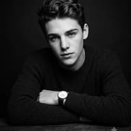 AI generated monochrome studio portrait of a young man with wavy hair, arms crossed on a table, wearing a ribbed sweater and wristwatch.