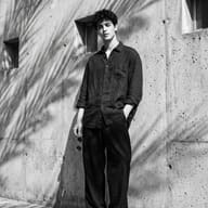 AI Image Prompt: Moody B&W fashion photography featuring a handsome man in an oversized shirt holding round sunglasses while leaning on a textured wall.