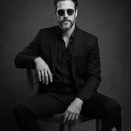 AI generated black and white studio portrait of a bearded man in a dark suit and sunglasses sitting on a wooden chair.