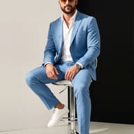 AI Image Prompt: High-end menswear studio photo of a bearded man in a sky-blue suit and sunglasses sitting on a chrome stool against a split black and white wall.