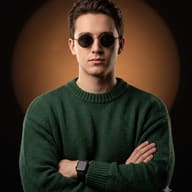 Man in Green Sweater and Sunglasses AI Photo Editing Prompt. AI generated studio portrait of a young man with curly brown hair and round sunglasses, wearing a forest green ribbed knit sweater, with crossed arms and a black smartwatch.