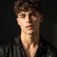 AI generated studio portrait of a curly-haired man in a black shirt holding a thin silver chain in his mouth against a dark background.