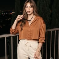 AI-generated fashion portrait of a woman in a rust satin button-up blouse and cream sweatpants, holding sunglasses to her lips while leaning on a railing against a blurred night city skyline.