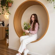 Cozy Cafe Lifestyle Photography AI Photo Editing Prompt. AI generated portrait of a young brunette woman in a pink sweater and white pants sitting in a circular wooden wall cutout, holding a bouquet of wildflowers inside a coffee shop.