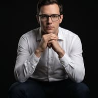 AI generated studio portrait of a man in a white shirt and glasses sitting with clasped hands against a black background.