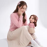 Realistic girl holding her matching 3D cartoon mini-me doll, both wearing pastel pink cardigans and beige pleated skirts.