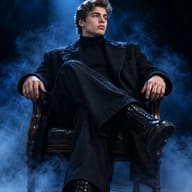 AI Image Prompt: Moody fashion photography of a handsome young man in a black turtleneck sitting elegantly in a dark room illuminated by a blue spotlight.
