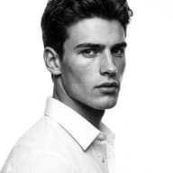 AI generated studio portrait photograph of a young man with detailed features and dark styled hair in a white shirt, looking forward against a clean white background.