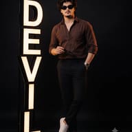 Fashion Studio Photography with 'DEVIL' Sign AI Photo Editing Prompt. AI generated studio portrait of a young man in a brown shirt and sunglasses standing next to a large illuminated vertical sign spelling 'DEVIL'.