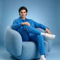 Man in a monochrome blue outfit and white sneakers sits in a plush, light blue textured armchair against a matching blue studio backdrop.