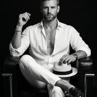 Man in White Linen on Leather Chair AI Photo Editing Prompt. AI generated monochrome studio portrait of a stylish bearded man in an open white linen shirt and trousers sitting on a black leather armchair while holding a fedora.