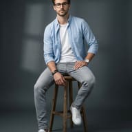 Young Man in Blue Shirt AI Photo Editing Prompt. AI generated studio portrait of a stylish young man wearing glasses and a light blue shirt, sitting on a wooden stool against a dark grey background.